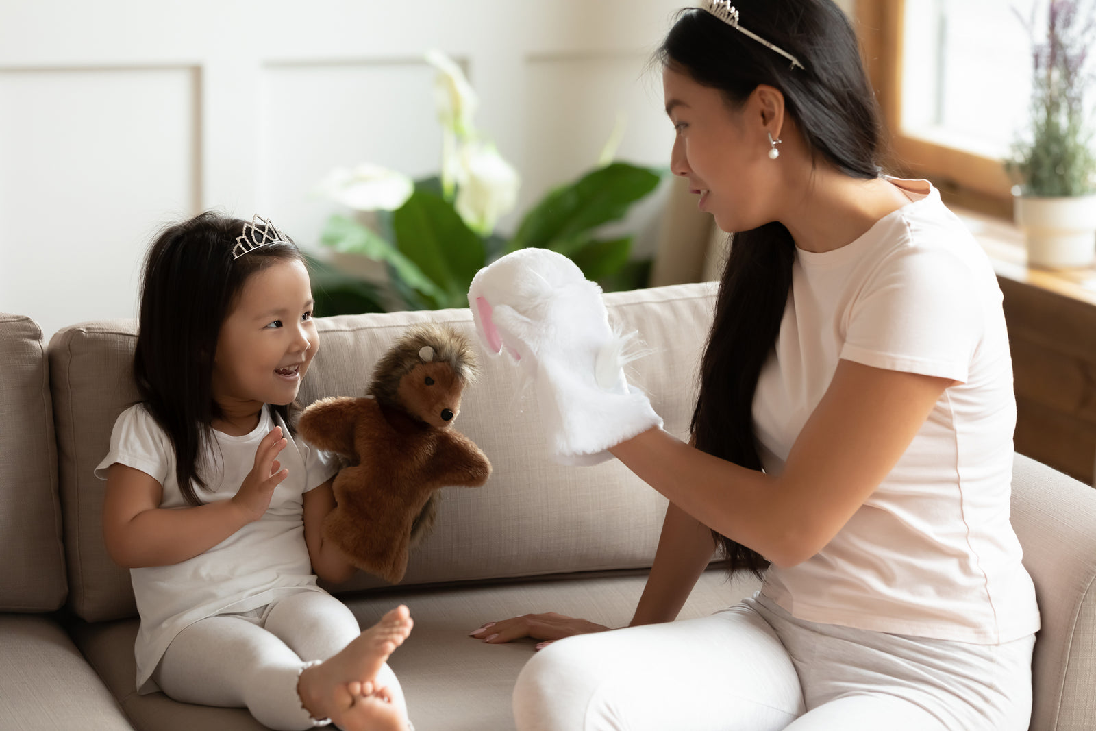 A mother and young daughter wearing tiaras, facing each other while seated on a couch, each with a puppet on their right hand.