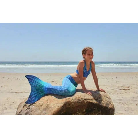 Front view of a young girl on a rock at a beach wearing the Blue Lagoon Mermaid Tail + Monofin Set.