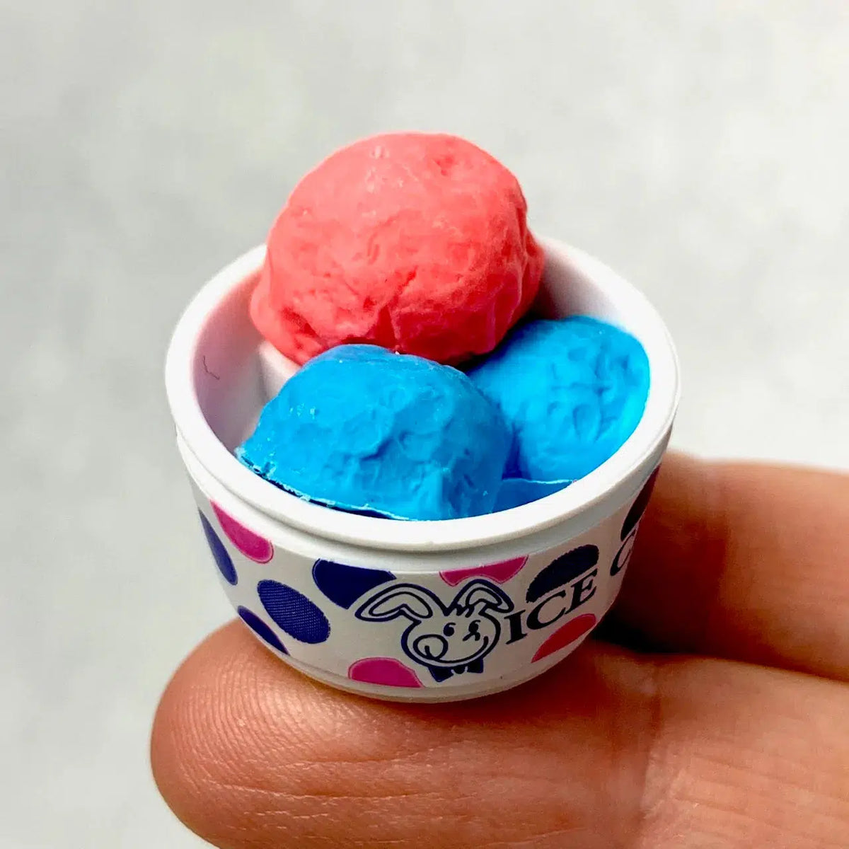 Front view of a small bowl of pink and blue Ice Cream Erasers sitting on a person&#39;s fingertips.