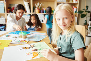 A girl looking directly at the camera, holding a paintbrush with a bright painted picture on the table in front of her. An adult and two children are in the background also working on a painting.