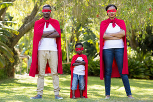 A father, young son, and mother standing in a line, facing the camera with their arms folded in front of them. They are all wearing red capes, tied at the neck and red masks covering their eyes.