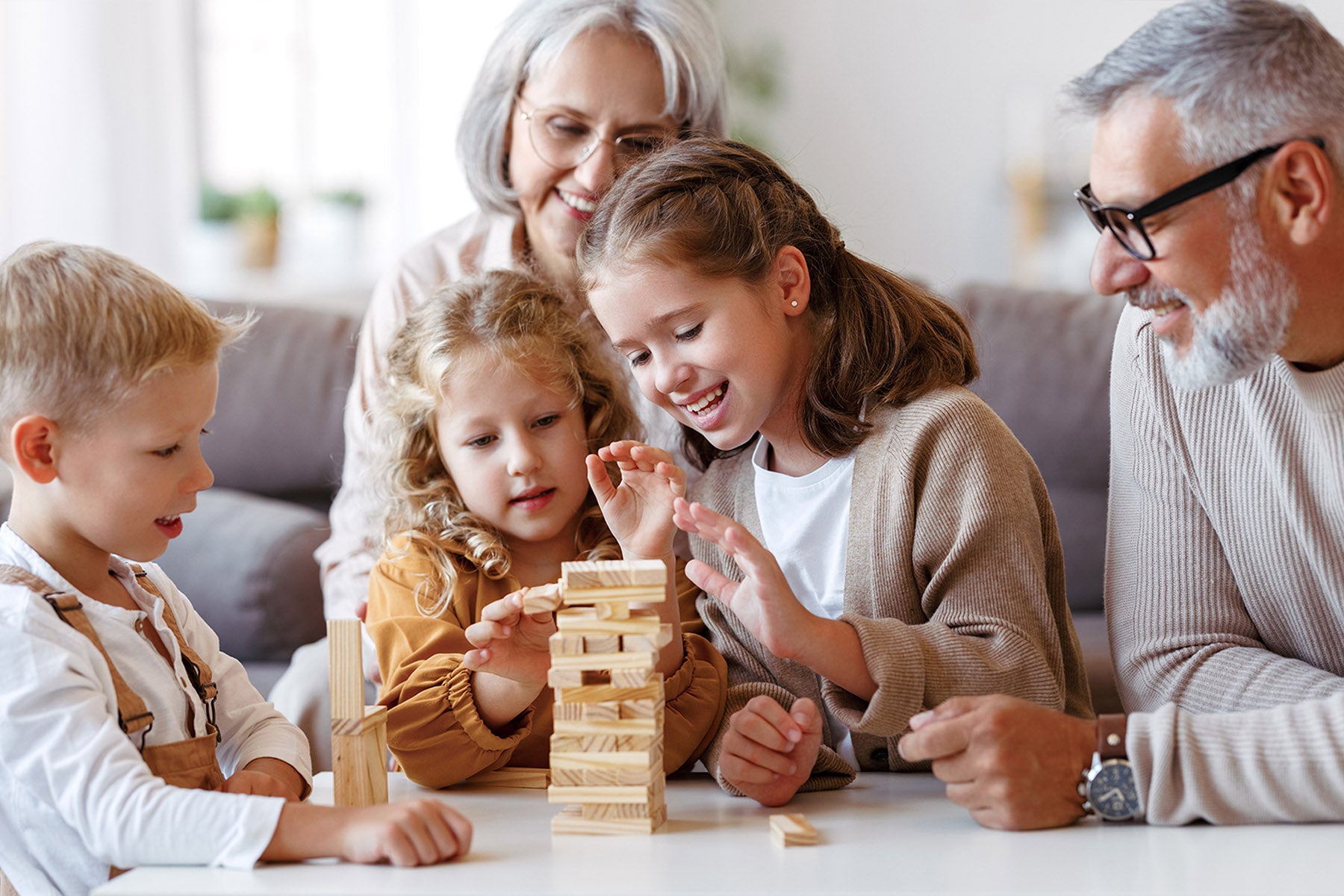Three children and their grandparents gathered around a coffee table, playing a stacking game made from natural wood pieces