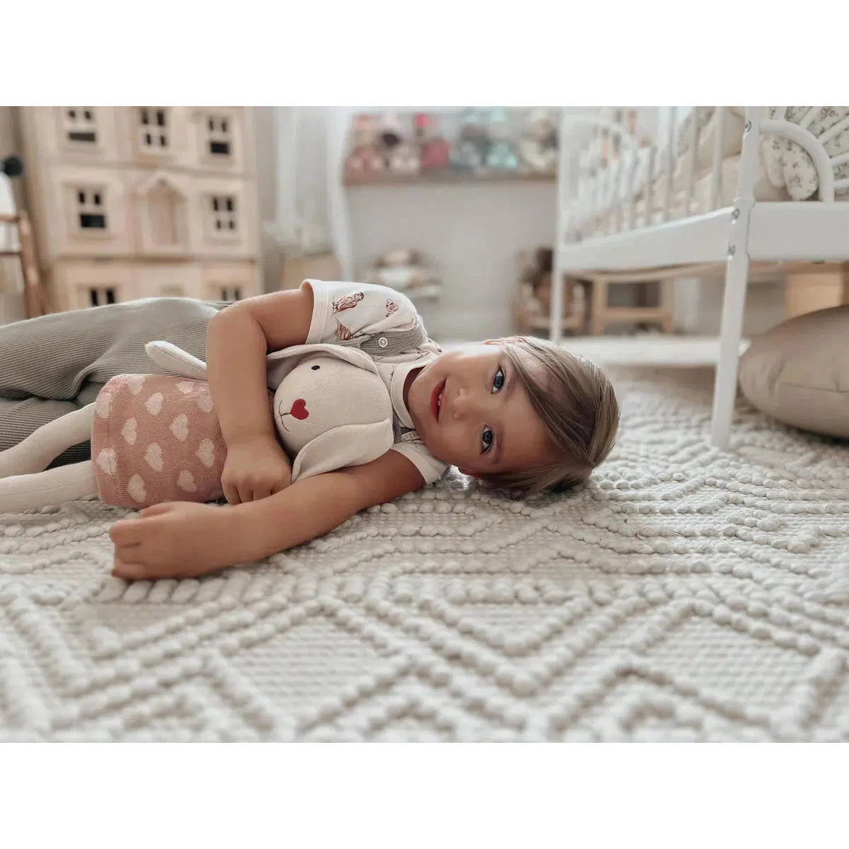 Front view of a young girl laying on the floor and holding Knit Bunny Plush-Luella.