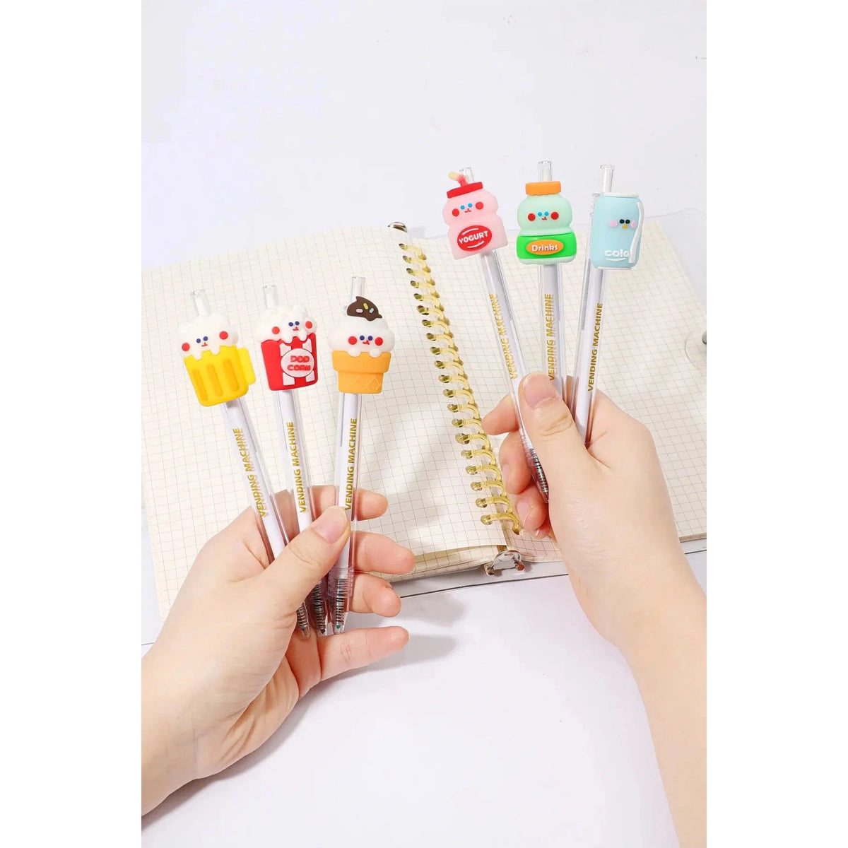 Front view of a person&#39;s hands holding the assorted styles of the 24-Hour Snack Shop Retractable Pen including the beer, drink, yogurt, ice cream cone, pop corn and cola over a spiral notebook.