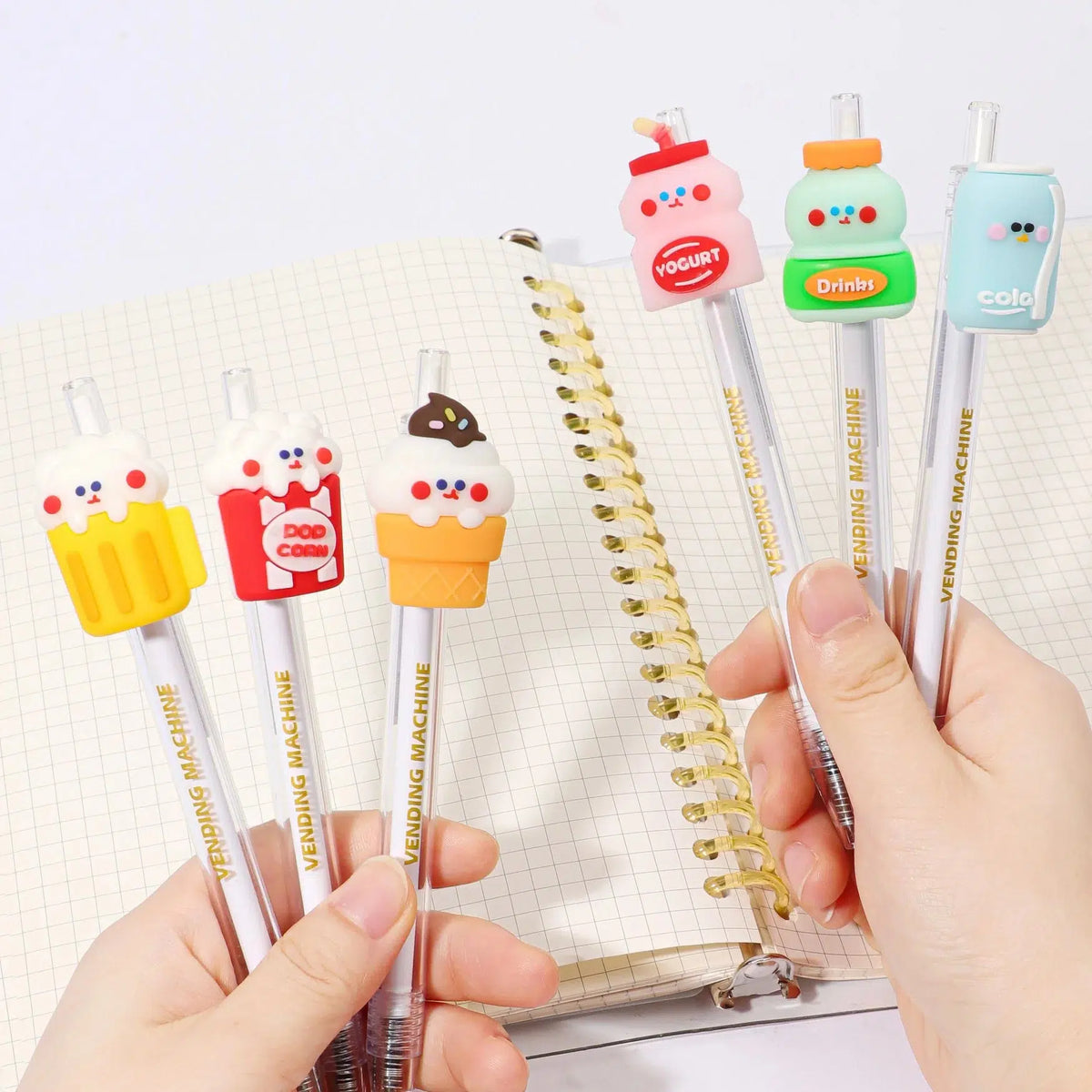 Front up close view of a person&#39;s hands holding the assorted styles of the 24-Hour Snack Shop Retractable Pen including the beer, drink, yogurt, ice cream cone, pop corn and cola over a spiral notebook.
