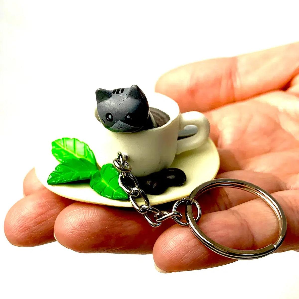 Front view of the Key Charm-Cat Cafe grey cat in a coffee cup being held in a person&#39;s hand.