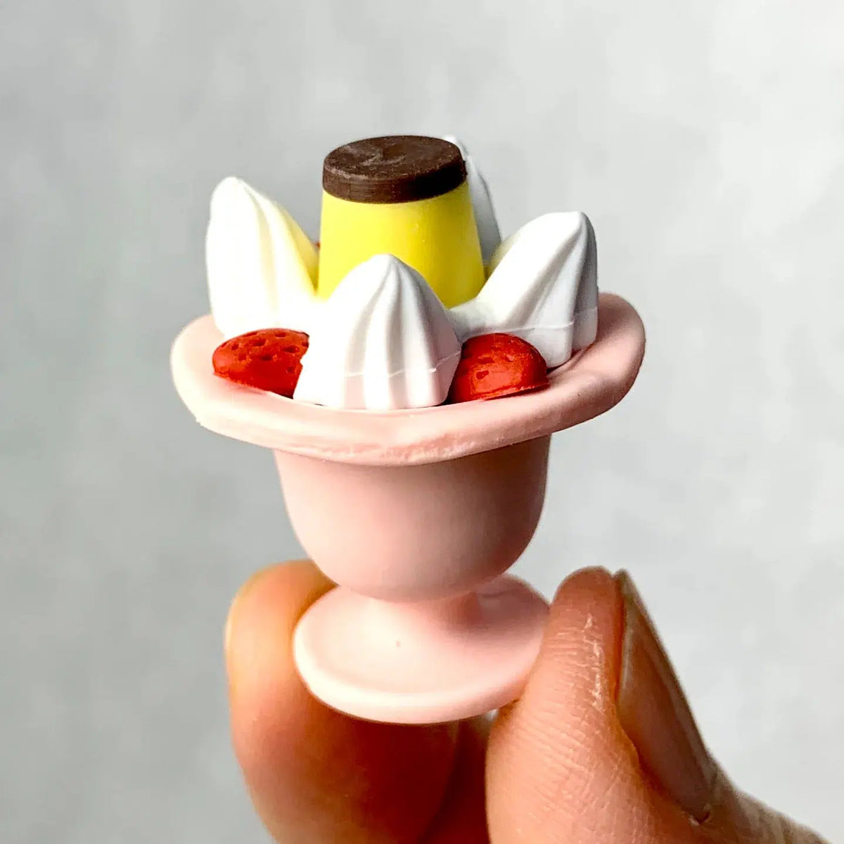 Front view of a person&#39;s fingers holding a pink sundae dish with strawberries, whip cream, and banana from the Puzzle Eraser Card Set-Dessert.