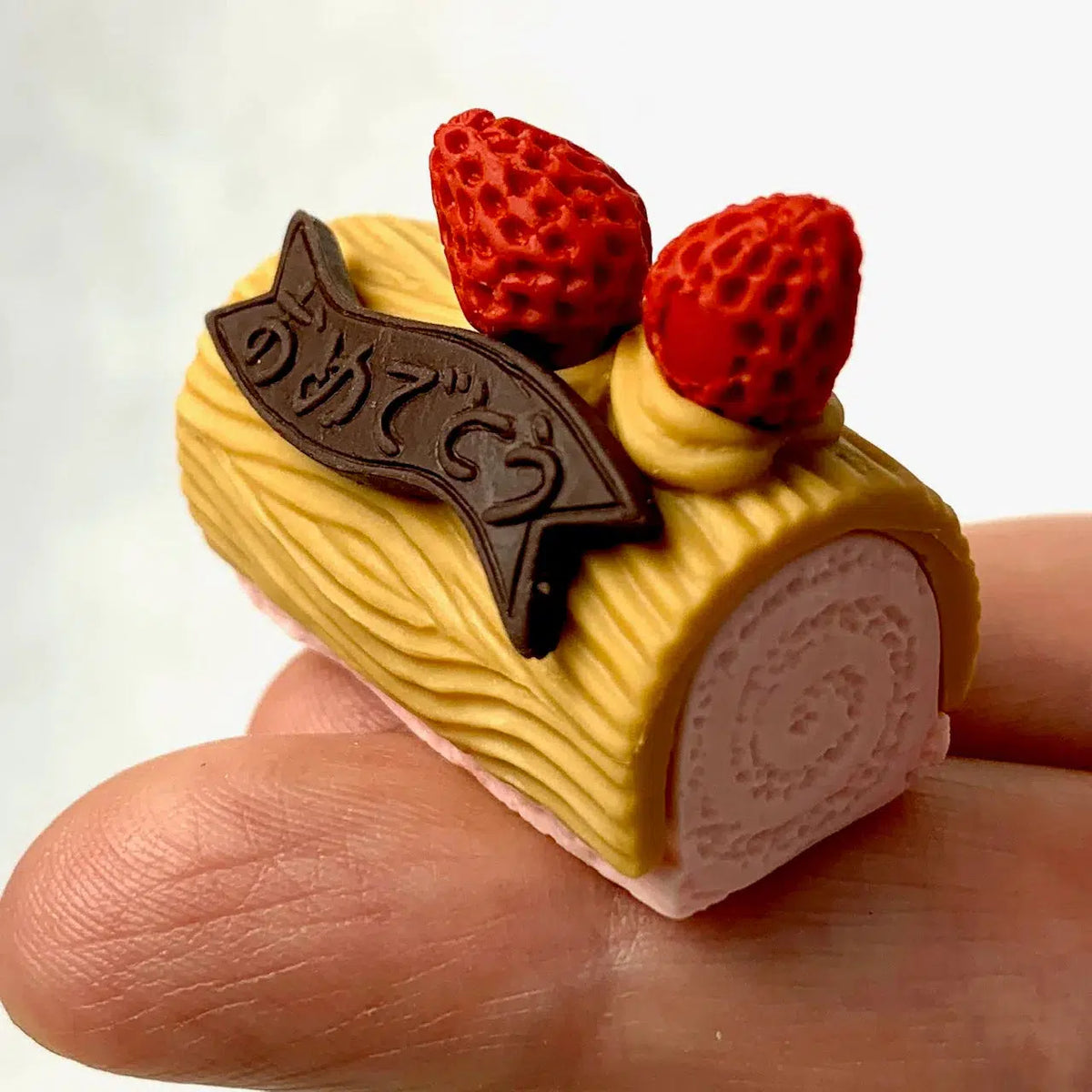 Front view of a person&#39;s fingers holding the pink log cake with yellow frosting, strawberries, and a chocolate banner from the Puzzle Eraser Card Set-Dessert.