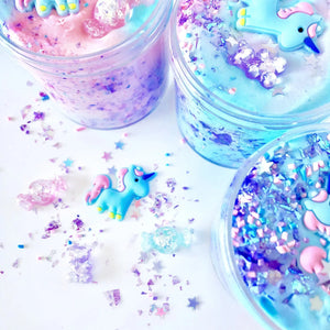 Front view of several containers of Don't Feed The Unicorns Butter Slime opened and decorated showing the glitter, candy pieces, and unicorn charm on table in front of them.