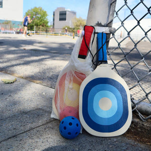 Front view of the Pickleball set laying against a fence showing the red paddle and ball from the Pickleball set in the mesh bag.