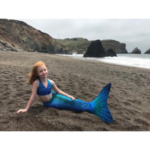 Front view of a young girl sitting on a Beach wearing the Blue Lagoon Mermaid Tail + Monofin Set.