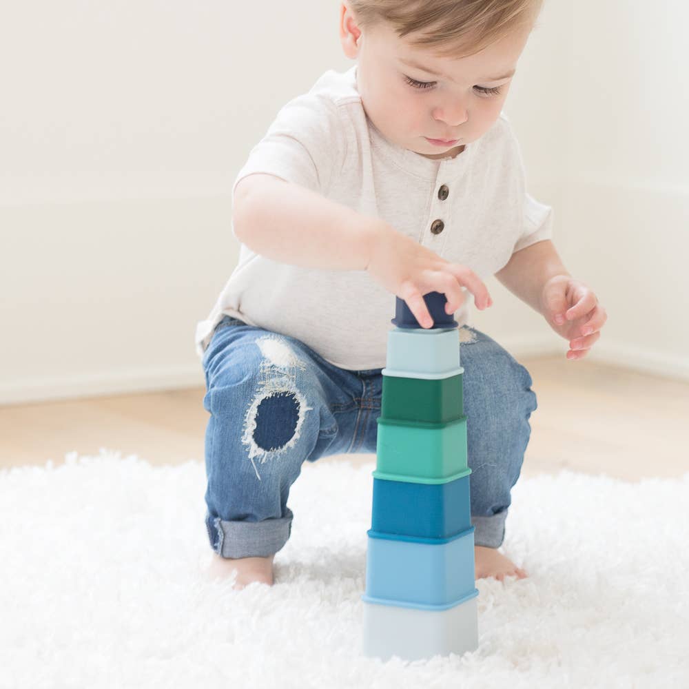 Front view of a baby squatting to place the top piece on his tower he made from Cool Blue Happy Stacks.