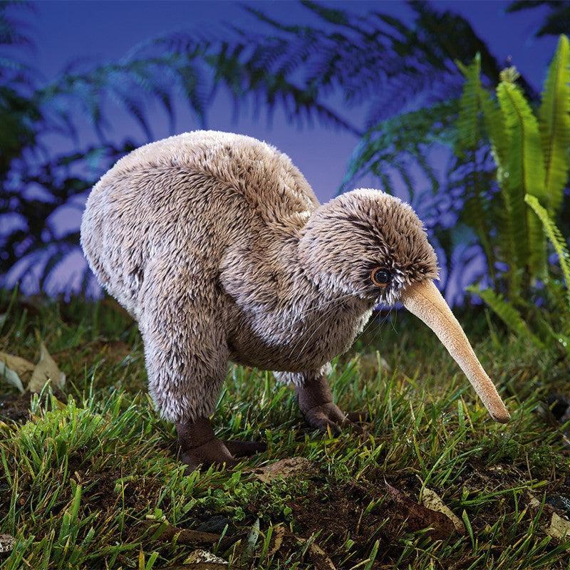 Plush toy of a kiwi bird standing on grass with a dark blue sky and ferns in the background