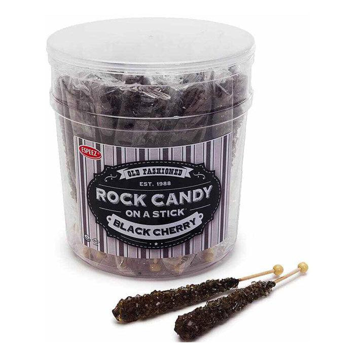 Front view of a display jar of black cherry rock candy. There are two rock candy sticks laying in front of the jar.