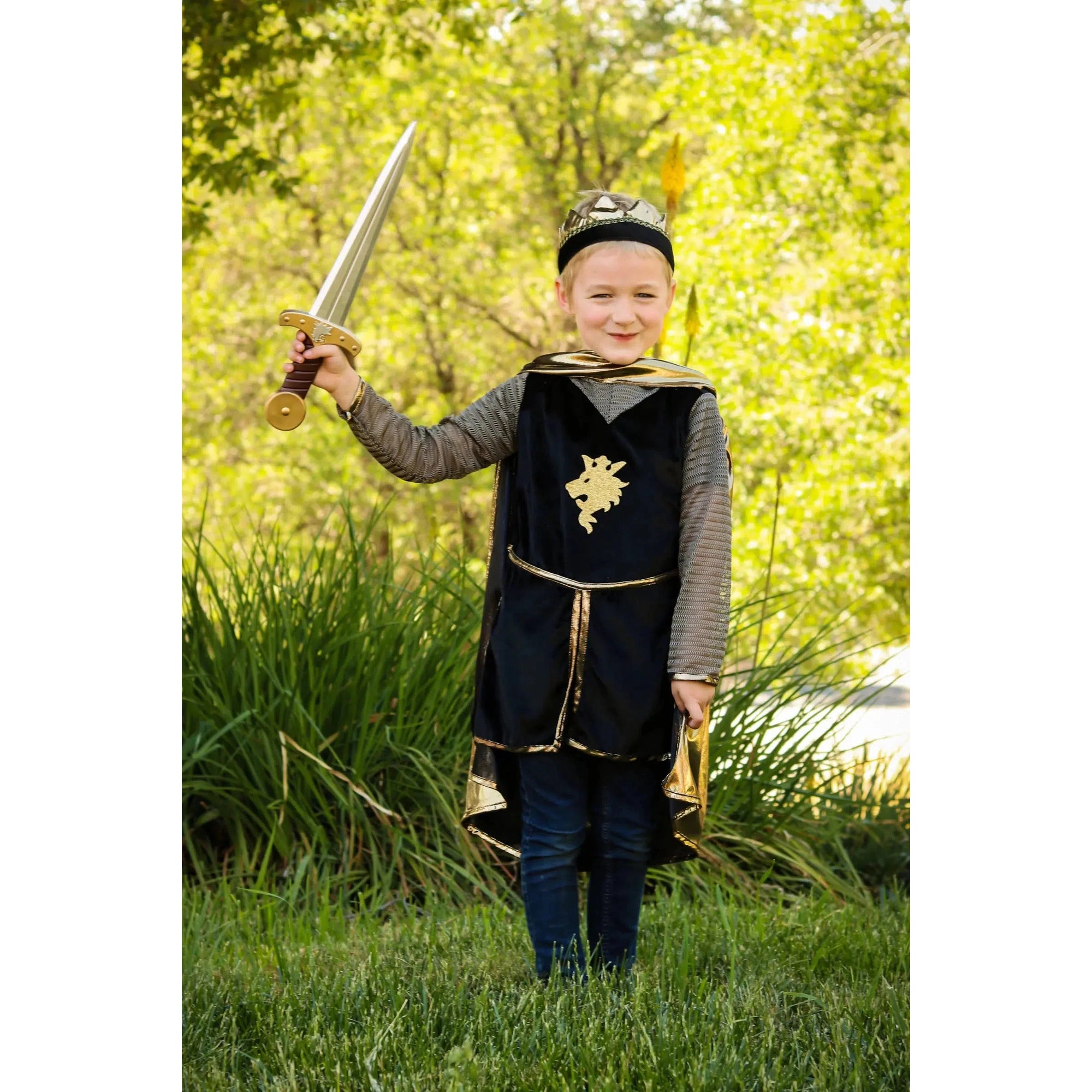Smiling child holding up a sword in his right hand, with the whole knight ensemble with the tunic, cape and crown.