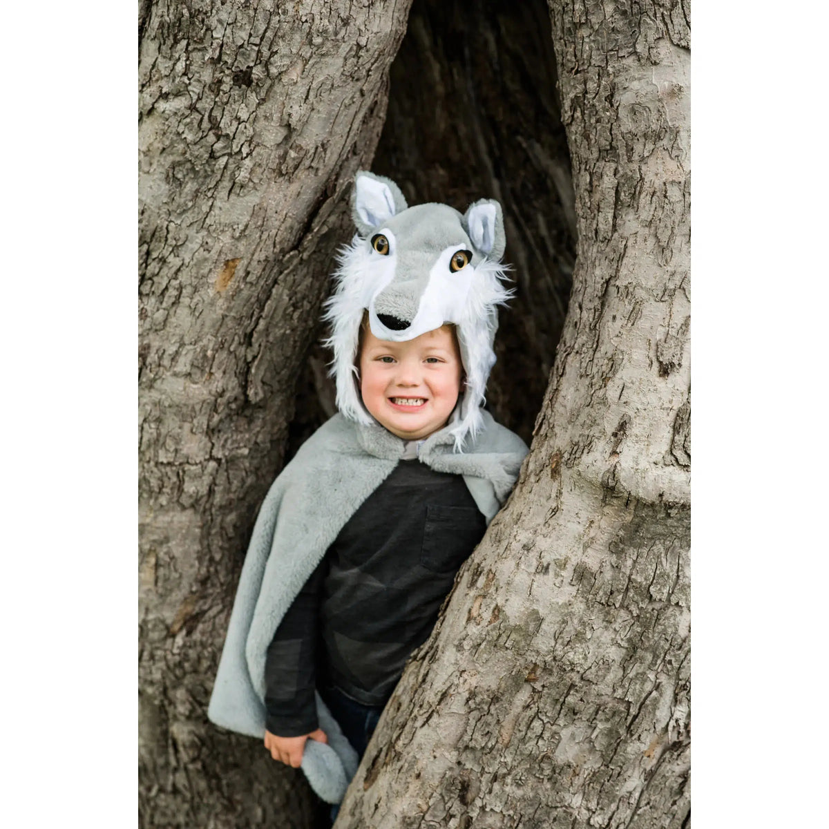 Child in woods, wearing a grey and white wolf cape, hiding in tree trunk