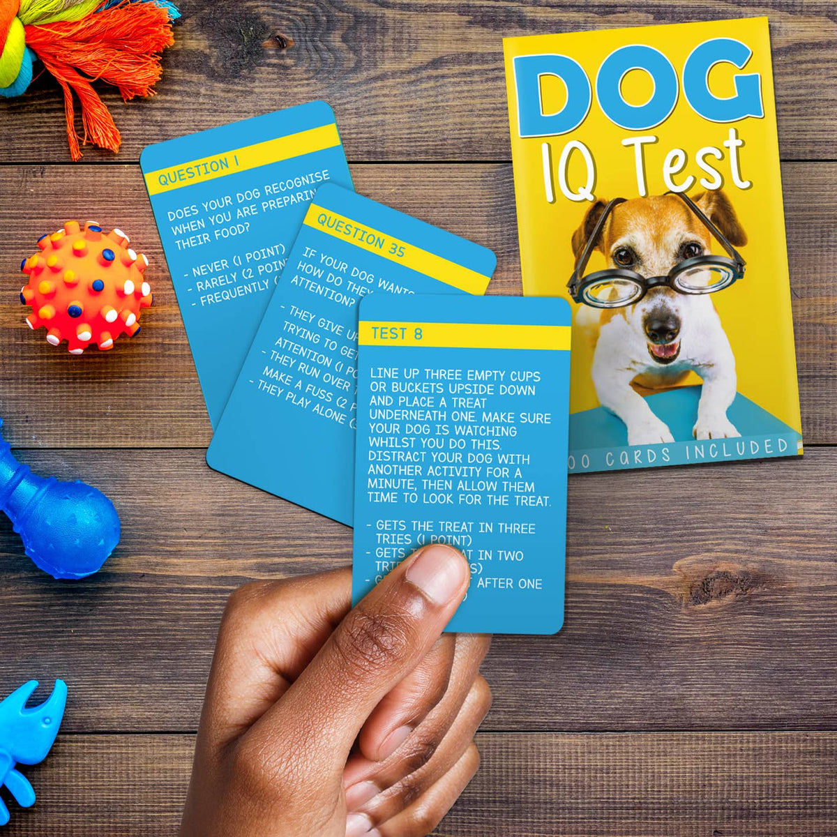 Front view of a persons hand holding a card included in the dog IQ deck. There are 2 other cards and the box to the deck in the background.