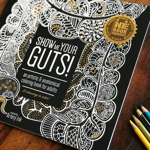 Front view of a copy of the adult coloring book Show Me Your Guts! on a table with four colored pencils beside it.