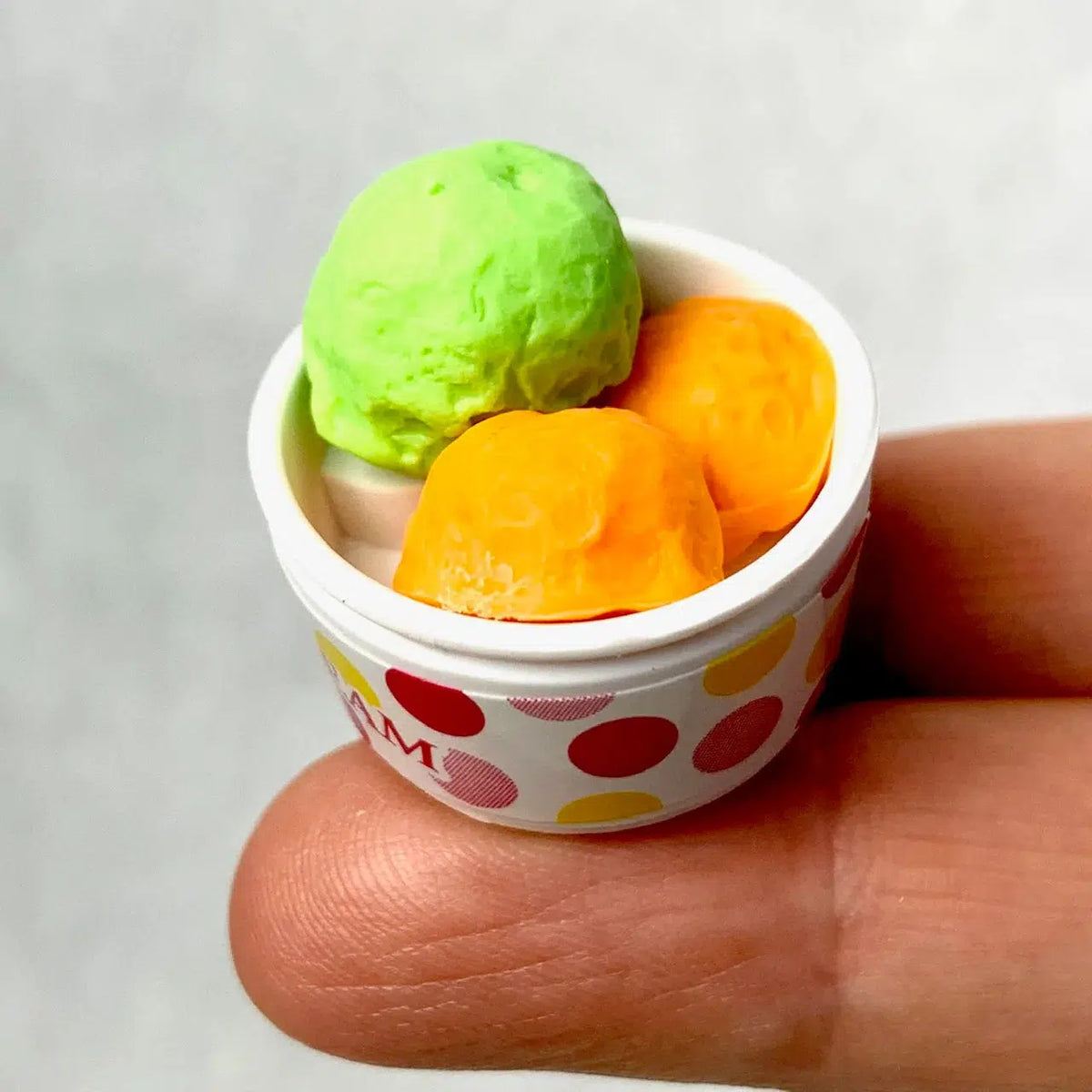 Front view of a green and orange ice cream in a bowl from Ice Cream Erasers sitting on a person&#39;s fingertips.