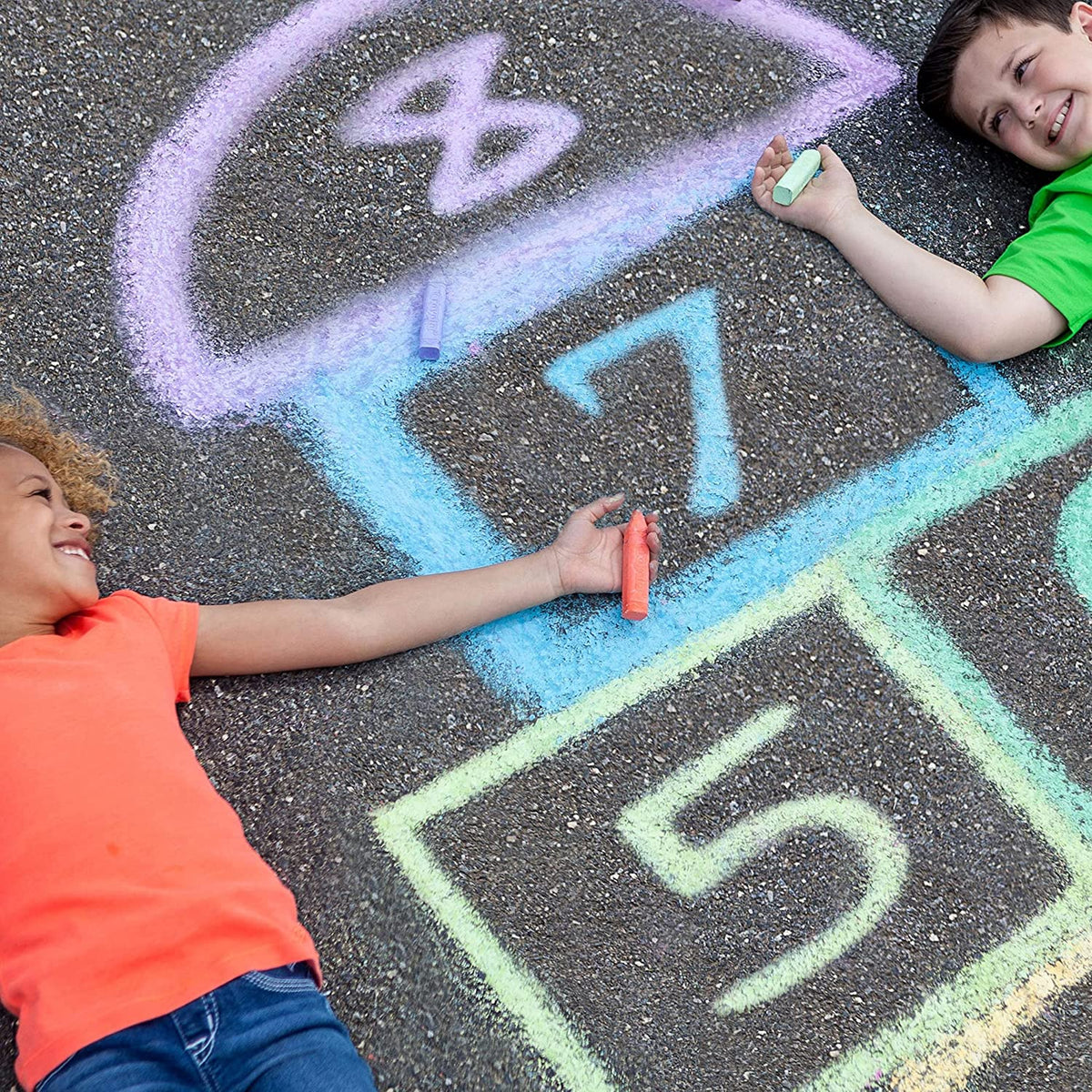Front view of two children laying on the ground on either side of a hopscotch both holding a Crayola 64 count Ultimate Sidewalk Chalk stick in their hands.