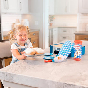 Front view of a young girl holding up a plate of the cinnamon rolls from the Prentendables Cinnamon Roll Set with the oven, mitt, can of rolls, plates, knife, and piping bag nearby.
