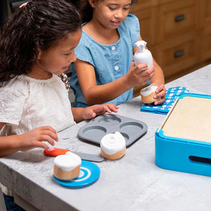 Front view of two girls playing with the Pretendables Cinnamon Roll Set with the muffin tin, oven mitt, knife, and plates and rolls nearby as one girl is using the piping bag to pretend to frost a roll.