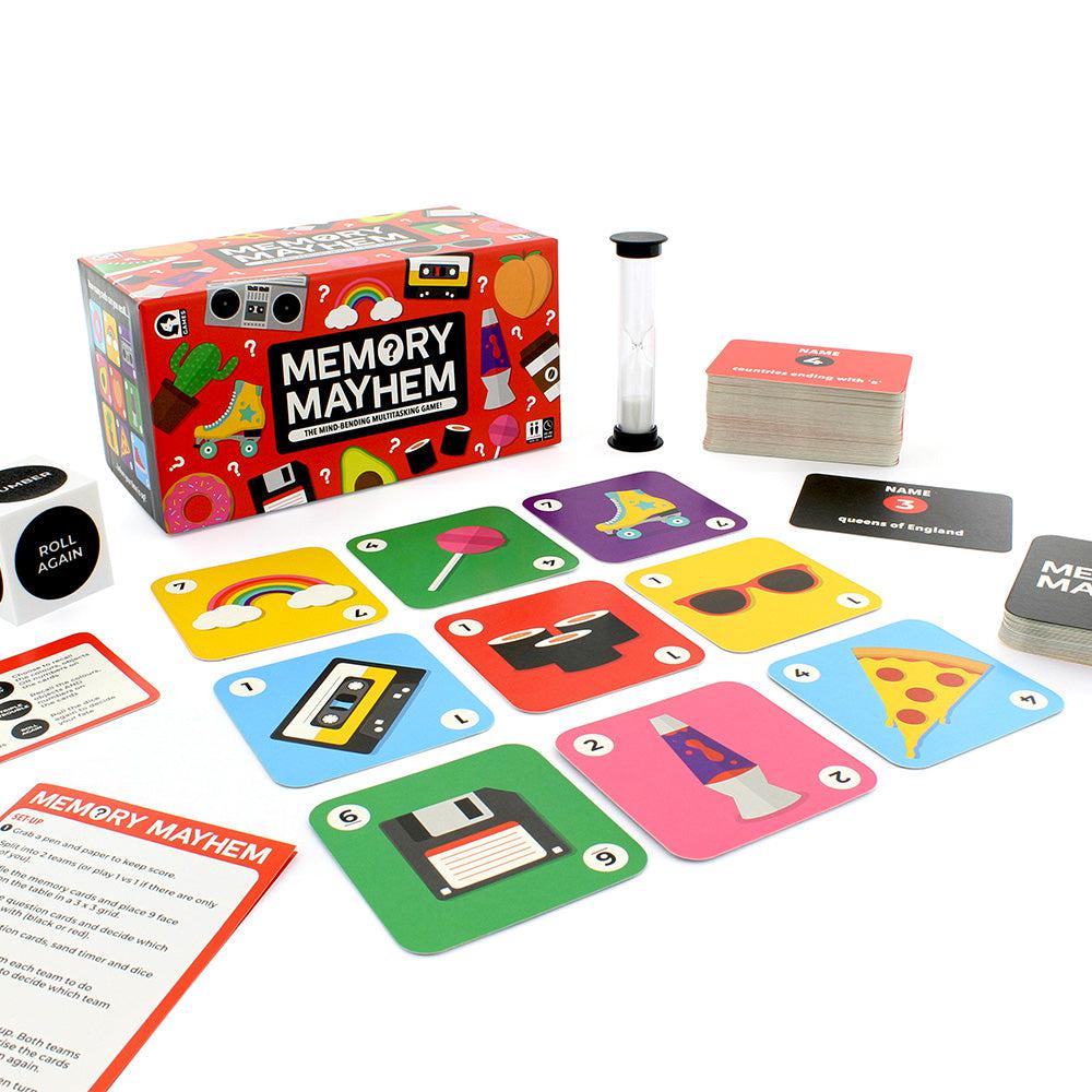 Front view of Memory Mayhem box with all the contents of game laid out in front and on the sides showing the timer, die, memory cards, and the questions for the memory game set up play.