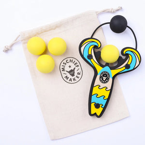 Front view of the yellow mischief maker slingshot and three yellow refill balls laying on bag it comes in.