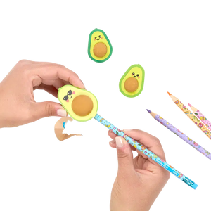 Front view of avocado sharpener being held in a person's hand with a pencil in it being sharpened while the avocado erasers lay beside with 3 pencils.