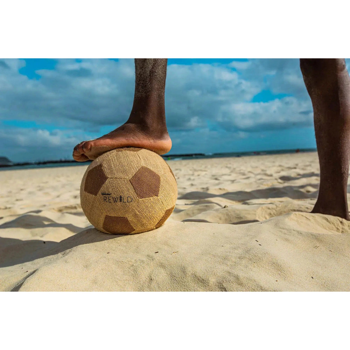 Front view of a beach with just the bottom half a person&#39;s legs with one foot on the Rewild Soccer Ball.