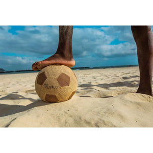Front view of a beach with just the bottom half a person's legs with one foot on the Rewild Soccer Ball.