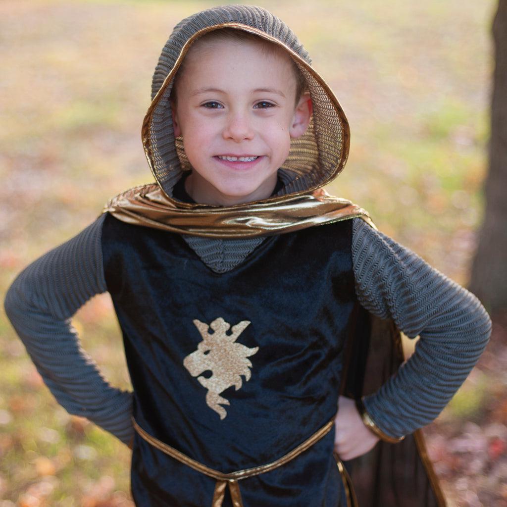 Smiling child holding up a sword in his right hand, with the whole knight ensemble with the tunic, cape and crown.