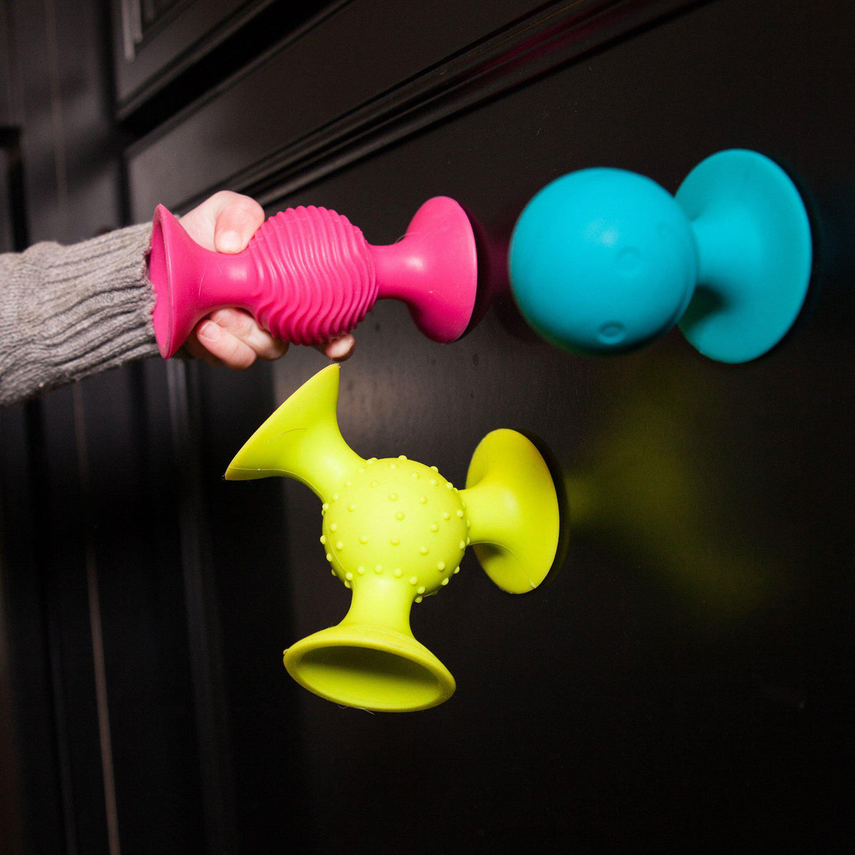 Colorful pipSquigs vertically suctioned onto black  cabinetry, with a child&#39;s hand grabbing the magenta one.