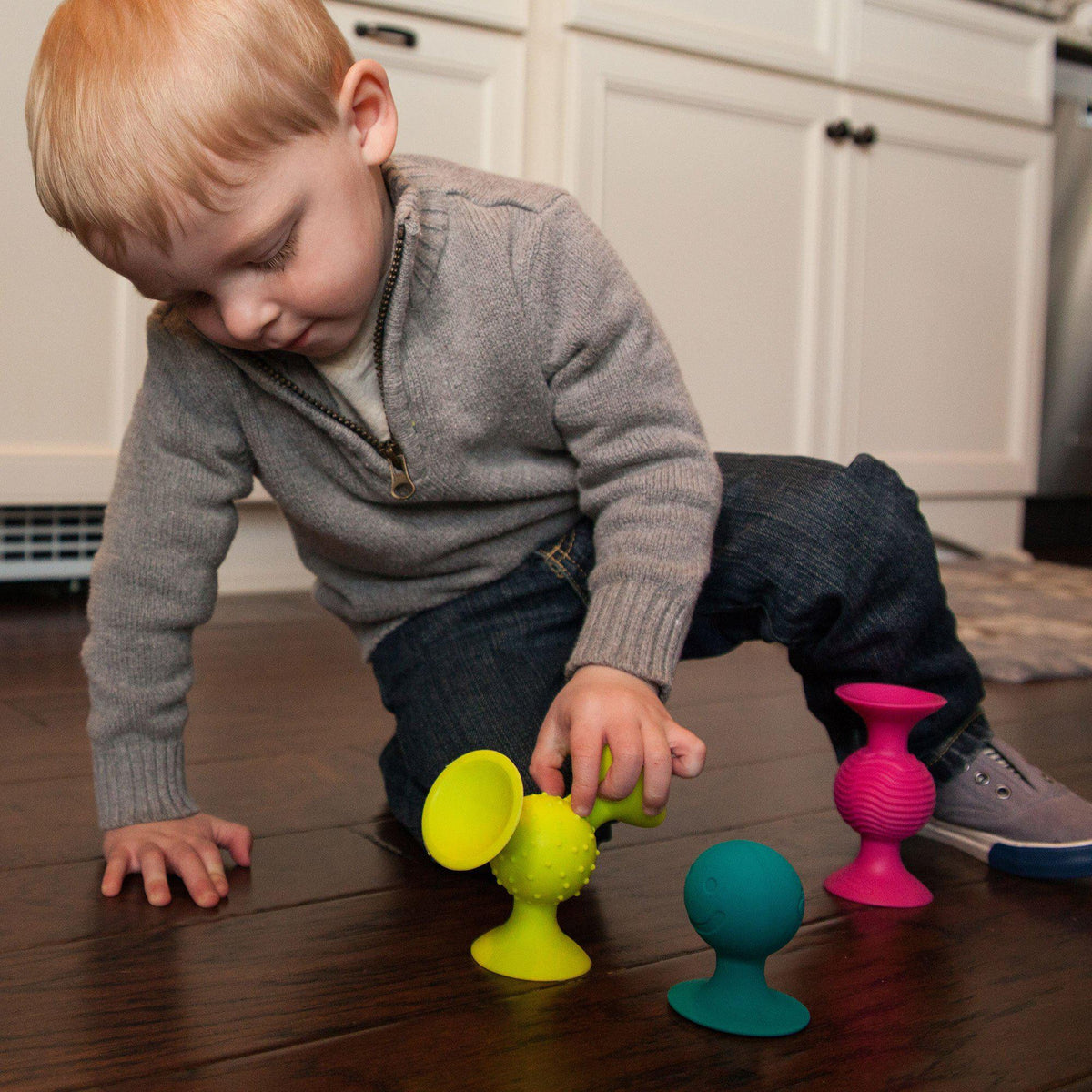 A red headed toddler playing with pipSquigs that are suctioned onto a wooden floor.